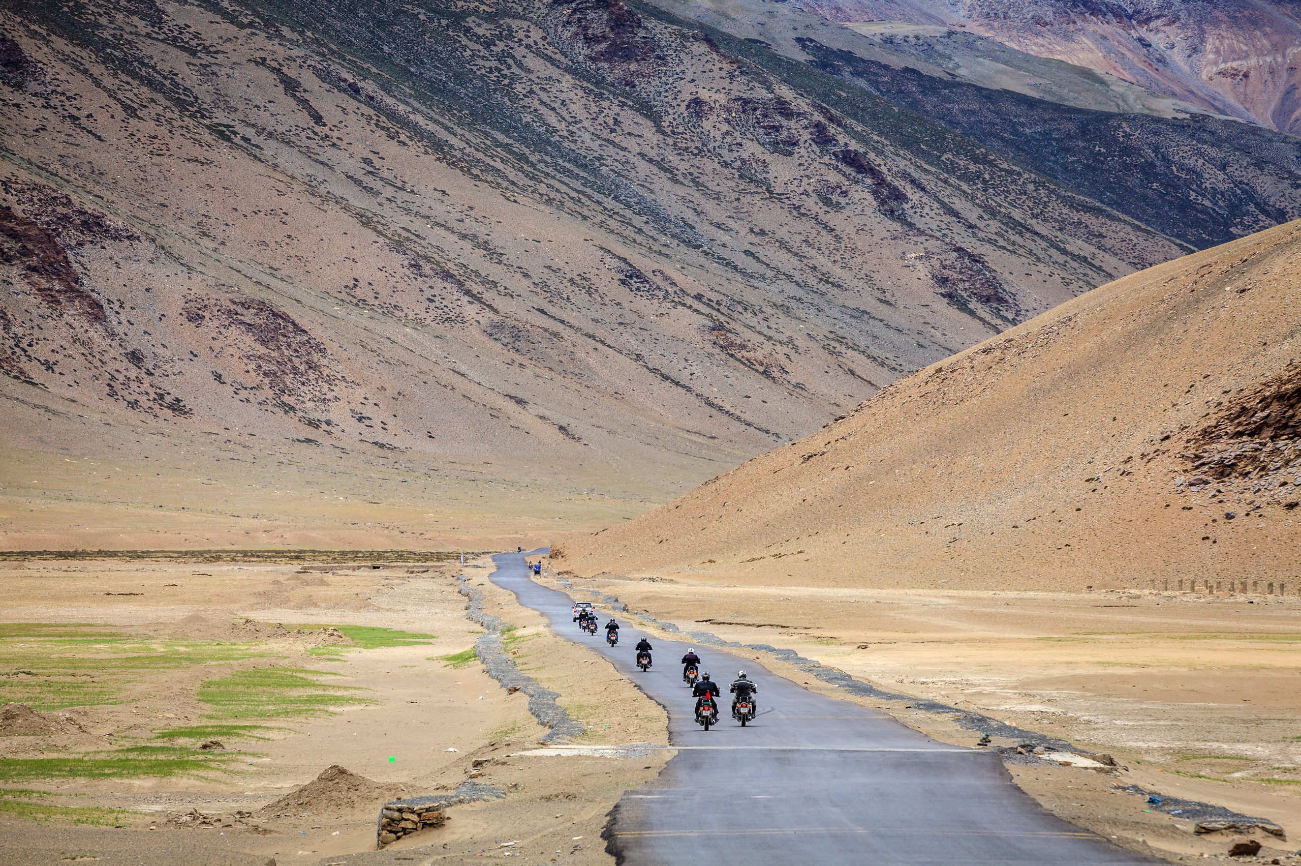 A line of motorbikes riding a narrow tarmac road through a wide Ladakh valley, bare brown mountains rising steeply on both sides