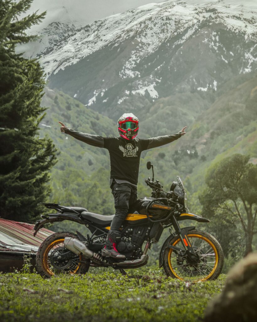 A rider standing on the pegs of a mud splattered Royal Enfield Himalayan 450, arms spread, with a snow covered mountain behind
